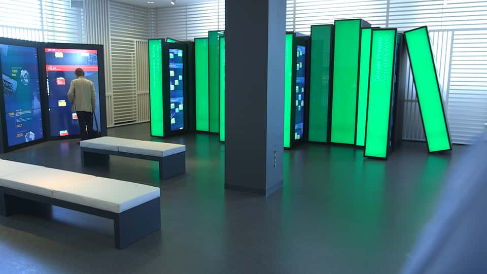 Large-scale interactive media installation at the BASF Visitor Center, showing an oversized digital book library with illuminated green vertical panels and integrated touchscreens, designed for spatial exploration of BASF’s corporate history.