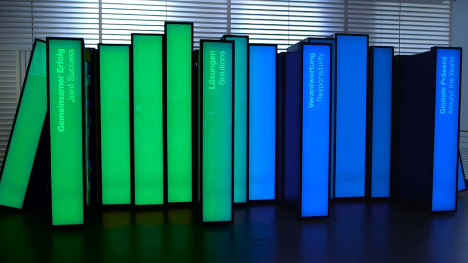 Wide view of the BASF History Library showing green and blue illuminated book panels, each representing a thematic chapter such as solutions, responsibility, and global presence.