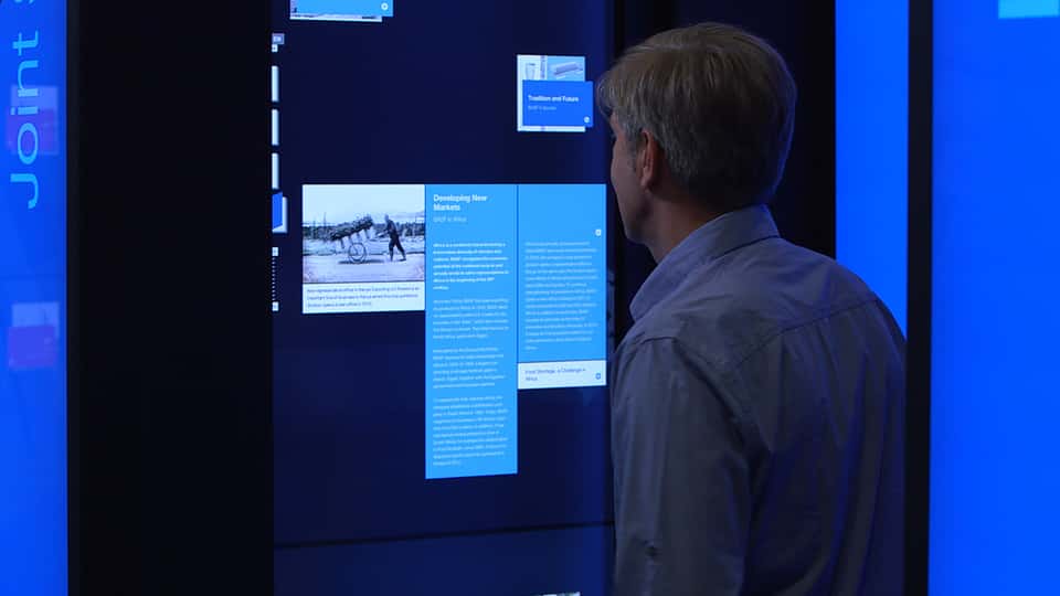 Visitor interacting with a vertical touchscreen displaying a data-driven historical timeline, part of the BASF History Library installation, featuring text, images, and archival content navigated through touch.