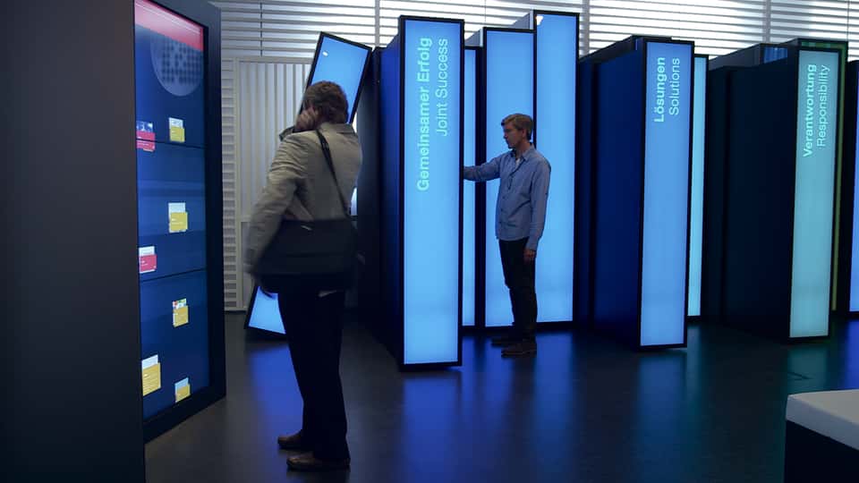 Visitors physically pulling and interacting with tall, book-shaped touchscreen panels in the BASF History Library, demonstrating the tangible interaction concept of the spatial media installation.