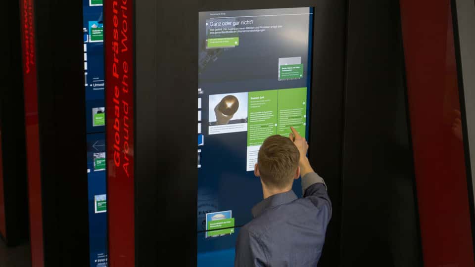Visitor using a vertical touchscreen embedded in a book-like installation element, exploring BASF historical content through touch-based navigation and data-driven text and media panels.