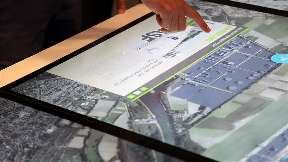 Visitor interacting with a large-format multitouch table displaying site data, images, and text panels within the BASF visitor center.