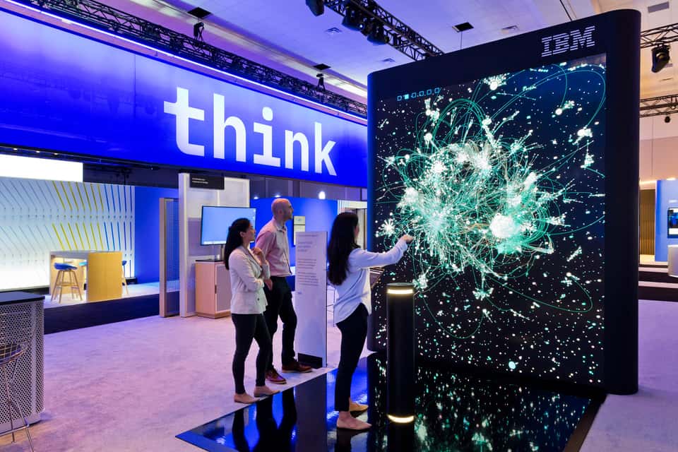 Photo of IBM booth at IBM Think 2023 showing the Under the Surface installation with a large, dark, 3D data visualization on a screen and people interacting with it