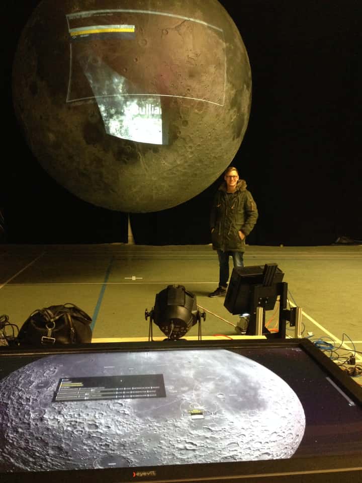 Claus Helfenschneider in front of a physically built moon model with live projection and the Moontable touch-enabled table in the background, showing a 3D visualization of the moon's surface with interactive elements.