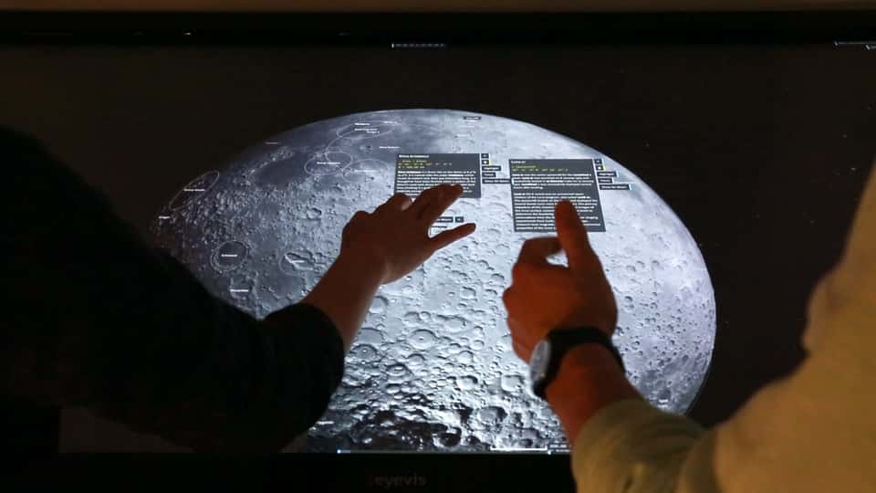 Photo of the Moontable installation showing a large multi-touch table with a 3D visualization of the moon's surface, with multiple users interacting with the display.